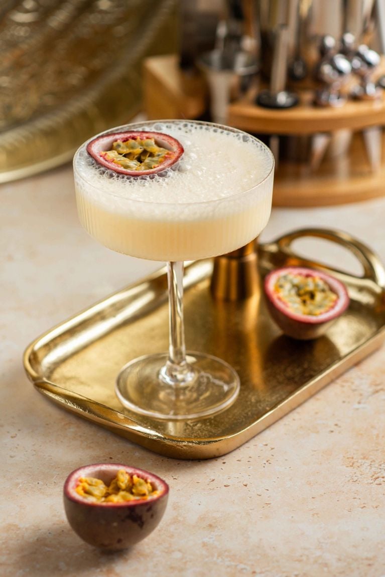 a passion fruit champagne cocktail cocktail is served in a coupe glass with a passion fruit half garnish floating on the foamy top layer, resting on a small, gold, rectangular serving tray. There are two additional halves of passion fruit on the tray and counter surface. The background is slightly blurred, showing bar equipment on a wooden stand.