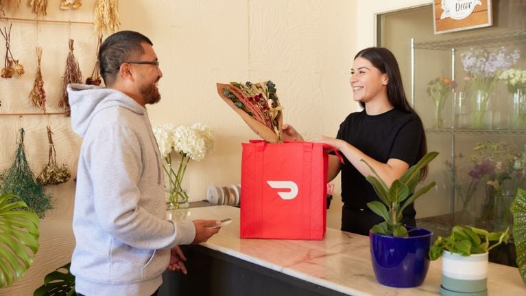 A man purchases flowers from a flower shop. A woman hands him his flowers with a red DoorDash bag on the counter.