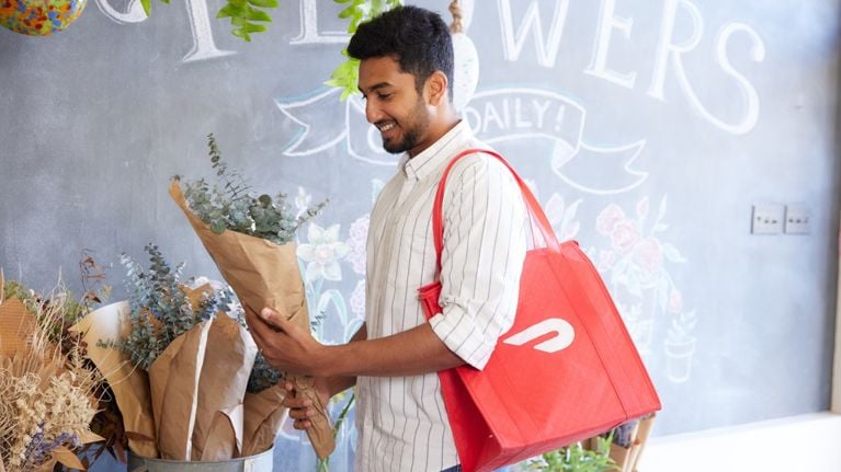 A man carries a bouquet of flowers with a red DoorDash bag on his shoulders.