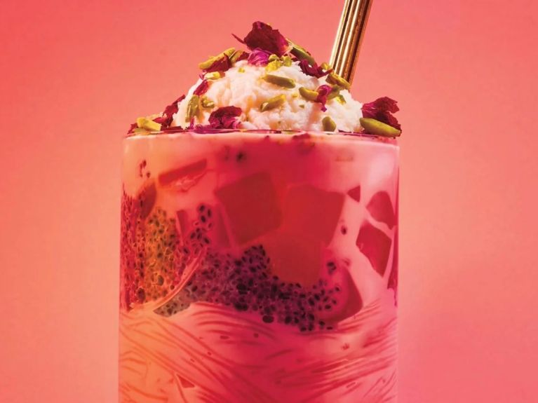 A tall highball glass filled with vermicelli noodles, sweet basil seeds, strawberry jello, ice cream, and pistachio rose petal garnishes.