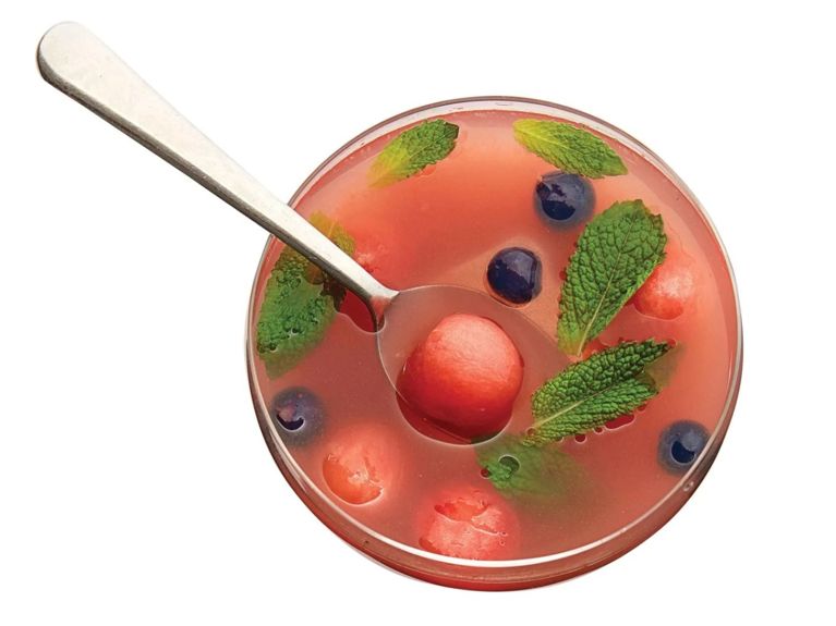 Chilled watermelon soup in a bowl with a spoon resting in it; fresh mint leaves and blueberries and watermelon balls float in it. The soup is made with watermelon; sugar; fresh mint; white wine; and blueberries for a post on a refreshing watermelon soup recipe for dessert