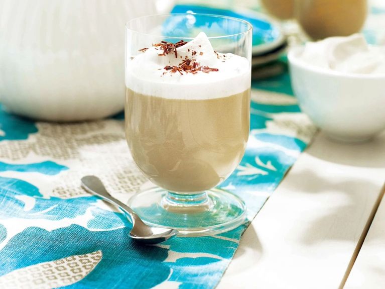 A dulce de leche pudding in a glass next to a spoon; the pudding is made with dulce de leech; whipping cream; sugar; cornstarch; milk and vanilla and garnished with whipped cream and shaved chocolate for a post on an easy dulce de leech pudding recipe
