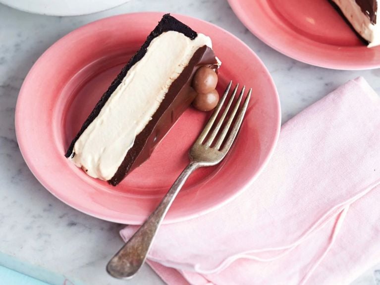 A pink plate with a slice of easy mocha ice cream cake topped with malteasers and coated with chocolate next to a fork and another slice of cake and a bouquet of pink flowers
