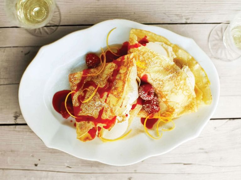 Frozen Dessert Crepes with vanilla ice cream topped with raspberries and raspberry sauce and orange peel on a white platter next to two goblets of white wine