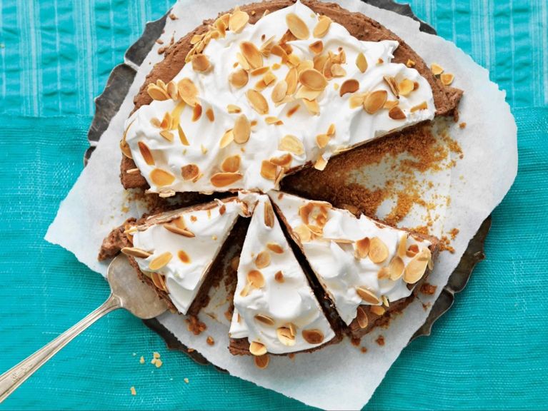 A frozen s'mores cake on a platter lined with parchment paper with a slice removed; the cake is made with graham-cracker crumbs; 35% cream; chopped bittersweet chocolate; and egg whites with a meringue topping topped with toasted sliced almonds for a post on a refreshing frozen s'mores cake recipe