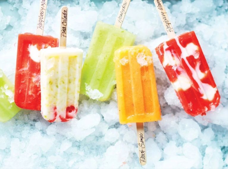 Green honeydew and lime paletas, orange Peach-Ginger Paletas, white and green Pina-Colada Paletas, and pink Raspberry And Mango Swirl Paletas with their names written on their popsicle sticks in cursive writing in an ice bucket for a recipe for four fresh fruit paletas
