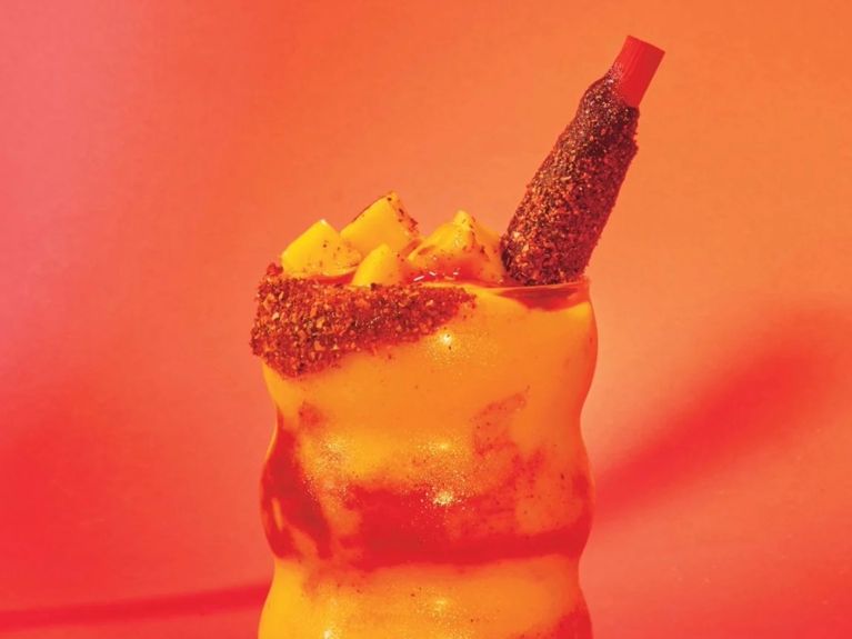 A wavy tall glass filled with mango slushy and stripes of chamoy sauce. It's dressed with chopped mango and a red straw.