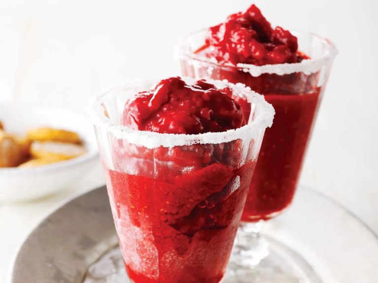 Two sundae glasses rimmed with sugar and salt filled with Raspberry-margarita sorbet on a serving tray filled with ice next to three spoons for a recipe for how to make Raspberry-margarita sorbet
