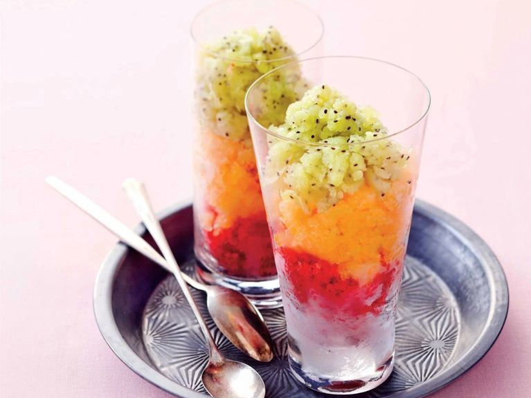Two strawberry-rhubarb granitas on a tray, the drinks are layered with green kiwi granita on top then orange granita then the strawberry rhubarb granita at the bottom with spoons next to them on a pink background