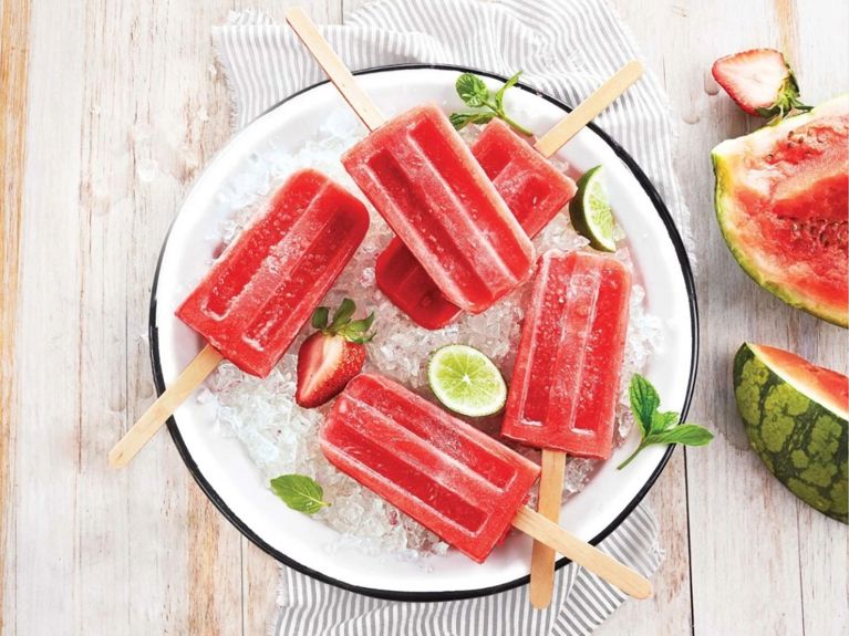 Strawberry-Watermelon Limoncello Ice Pops on a white plate filled with crushed ice next to slices of lime and fresh strawberries and fresh mint on a striped napkin on a wood table next to a cut watermelon and more strawberries