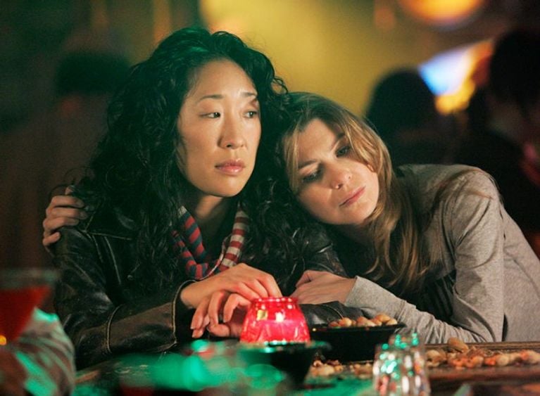 Two characters from Grey's Anatomy lean on each other at a bar