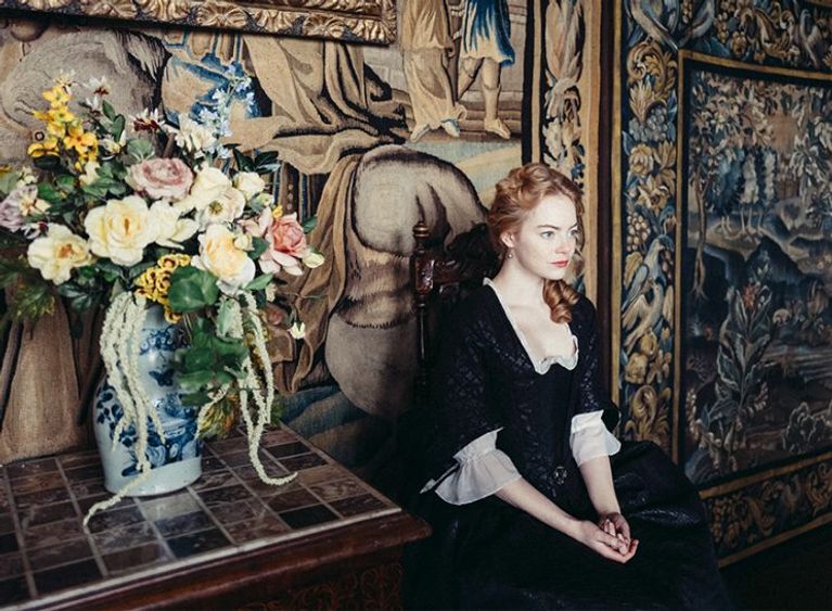 Emma Stone sits in a period costume against an ornate wall in a movie about Queen Anne called The Favourite