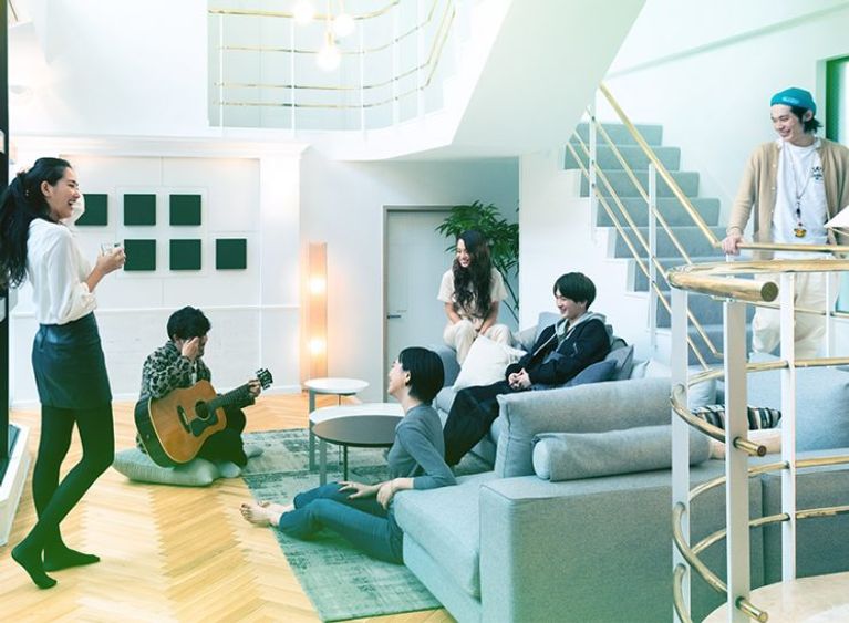 A few young people sit around on a grey couch as one of them plays the guitar