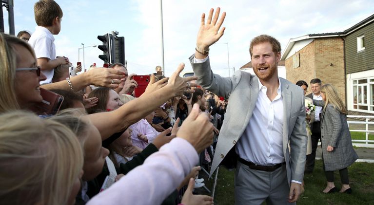 Prince Harry wearing a grey suit and white shirt waves to a crowd of fans