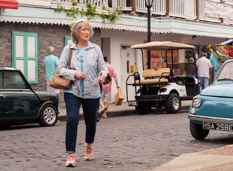 Meryl Streep wearing jeans, windbreaker and pink sneakers walks across a cobblestone street
