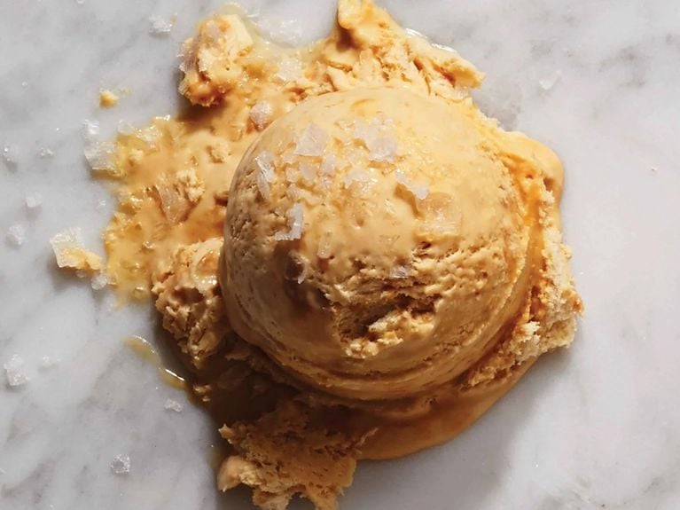 A single, large scoop of golden-tan salted caramel ice cream sitting on a white marble surface. The ice cream is slightly melting at the base and topped with large, translucent flakes of sea salt.