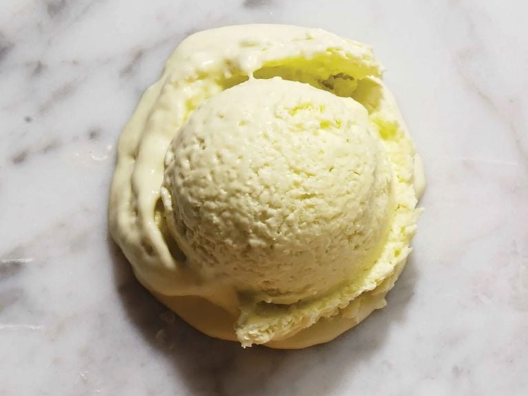 A top-down view of a single scoop of pale, pastel green-yellow avocado ice cream resting directly on a white marble surface. The scoop has a textured, creamy surface and is melting slightly, forming a thick, liquid pool around the base.