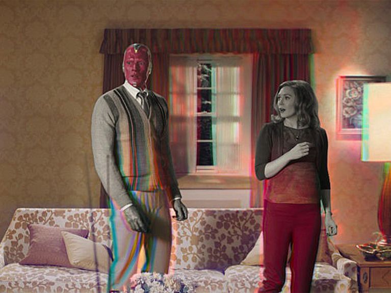 Man with a red, supernatural face stands next to a woman in a living room.