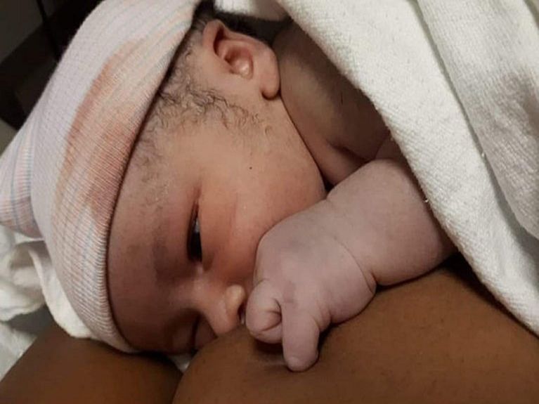 A newborn baby wearing a light pink hat and draped in a blanket lies on it's mother's chest