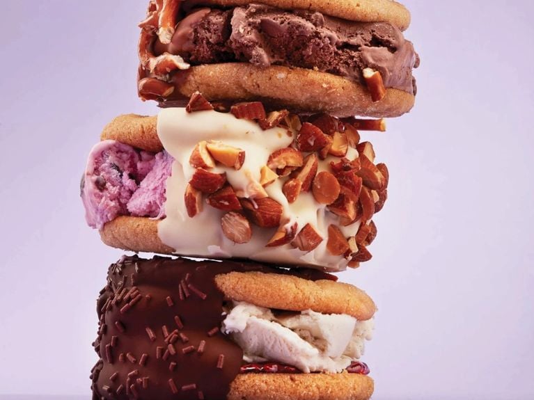 A stack of peanut butter cookie ice cream sandwiches rolled in chopped toasted pecans; some are filled with strawberry ice cream' some chocolate, half are dipped in white chocolate and half dark chocolate for a post on the best ice cream sandwich recipes