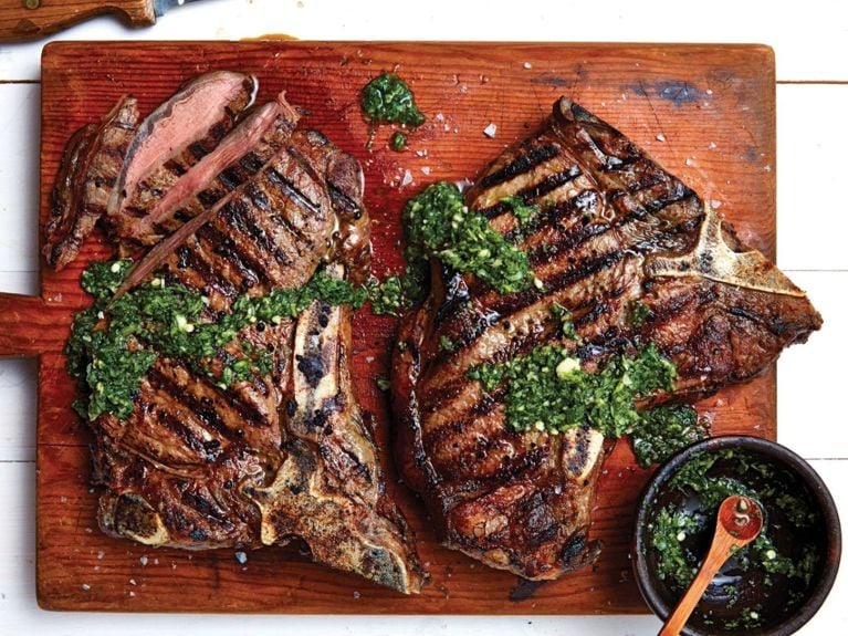 Classic steaks topped with green spicy Thai sauce on a wood cutting board next to a bowl of the sauce. The steaks are made with 4 T-bone steaks; the sauce is made of garlic; serrano chilies; lime juice; fish sauce; brown sugar; fresh cilantro and fresh mint for a post on an easy grilled classic steak recipe with spicy Thai sauce