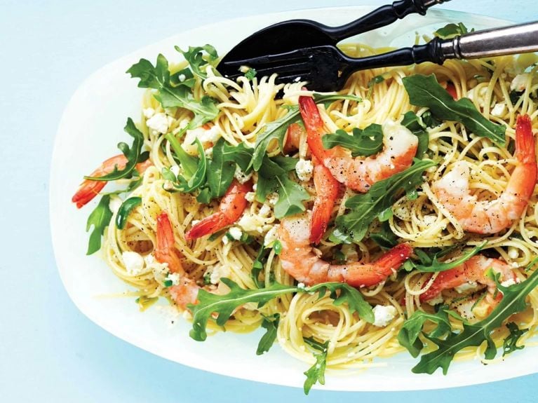 A white serving plate of linguine pasta covered with feta and shrimp and arugula with two black and silver serving tongs resting on it