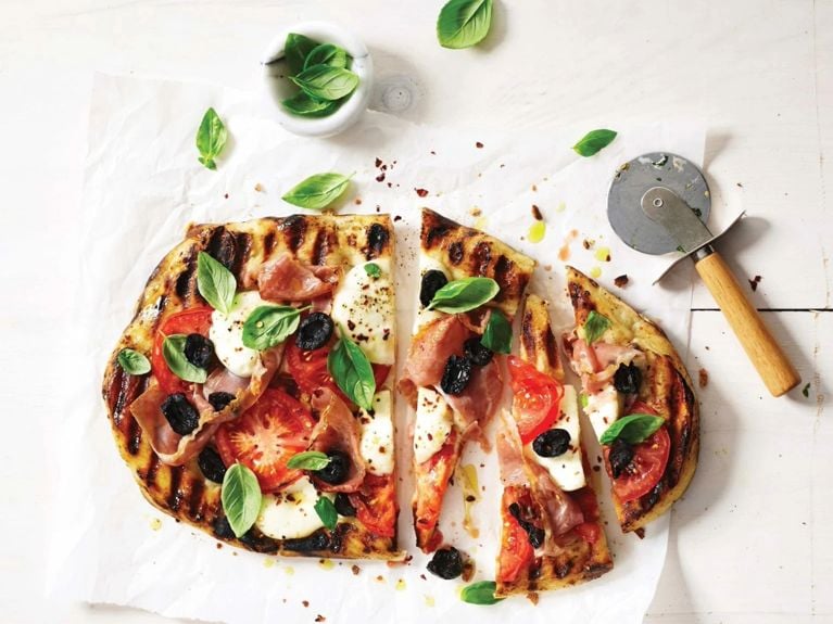 A grilled pizza with dark brown grill marks topped with fresh basil; tomato; mozzarella; pitted sun-dried black olives; chili flakes and prosciutto on parchment paper next to a pizza wheel for a post on an easy grilled pizza recipe