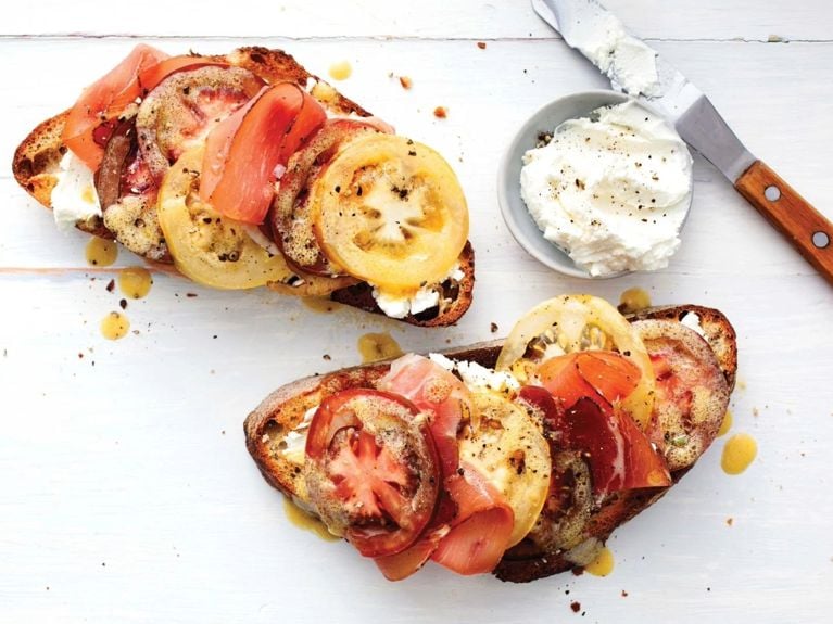 a tomato and prosciutto open-faced sandwich on rustic bread next to a small bowl of goat cheese with a spreading knife; the sandwiches are made with red beefsteak tomato, sliced into 1/4-in. rounds; yellow beefsteak tomatoes; white balsamic vinegar; olive oil; honey; dijon mustard; garlic; sourdough bread; goat cheese; prosciutto and pepper for a simple open-faced summer tomato sandwich recipe