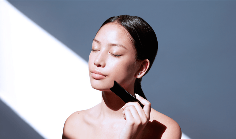 A woman using a gua sha tool against a bright background.