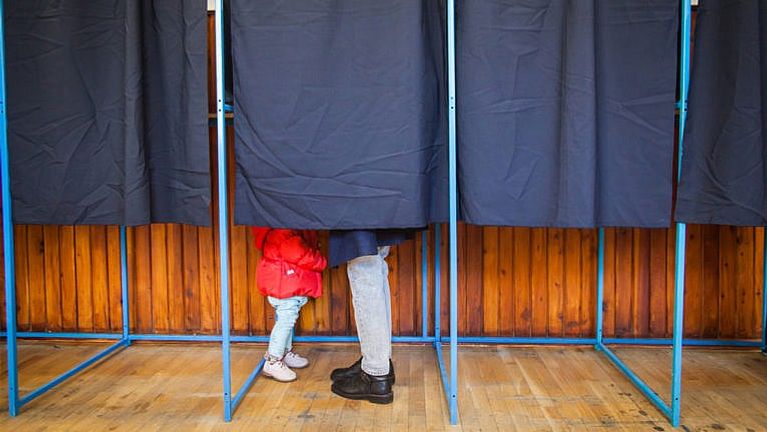 A photo of a curtained voting booth, with the feet of a small child in a red coat and a parent sticking out under the curtain