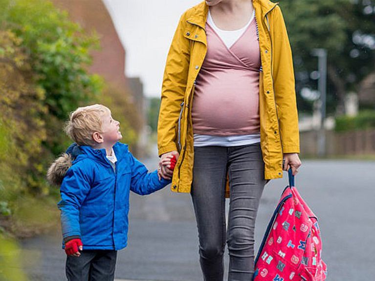 Child wearing a blue winter coat holding their pregnant mother's hand walking to school