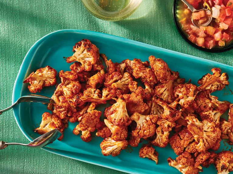 A green platter on a teal tablecloth holding charred cauliflower with tongs resting on the side