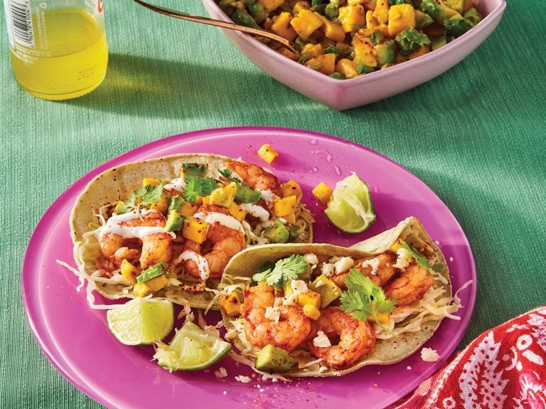 A pink plate with tacos filled with smoky chipotle and chiles de arbor shrimp with lime wedges on a teal tablecloth beside a salad and a turned bottle of jarritos