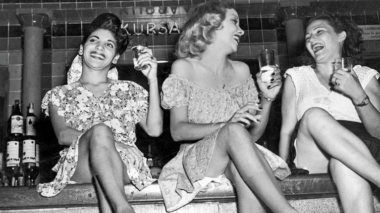 A black and white photo of three young women in dresses, sitting on a bar, holding cocktails and having a great time