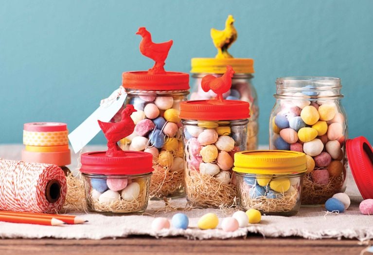 Five glass jars of varying sizes filled with pastel-colored candy eggs and straw, featuring lids topped with small plastic chicken figurines in red and yellow, set against a teal background.