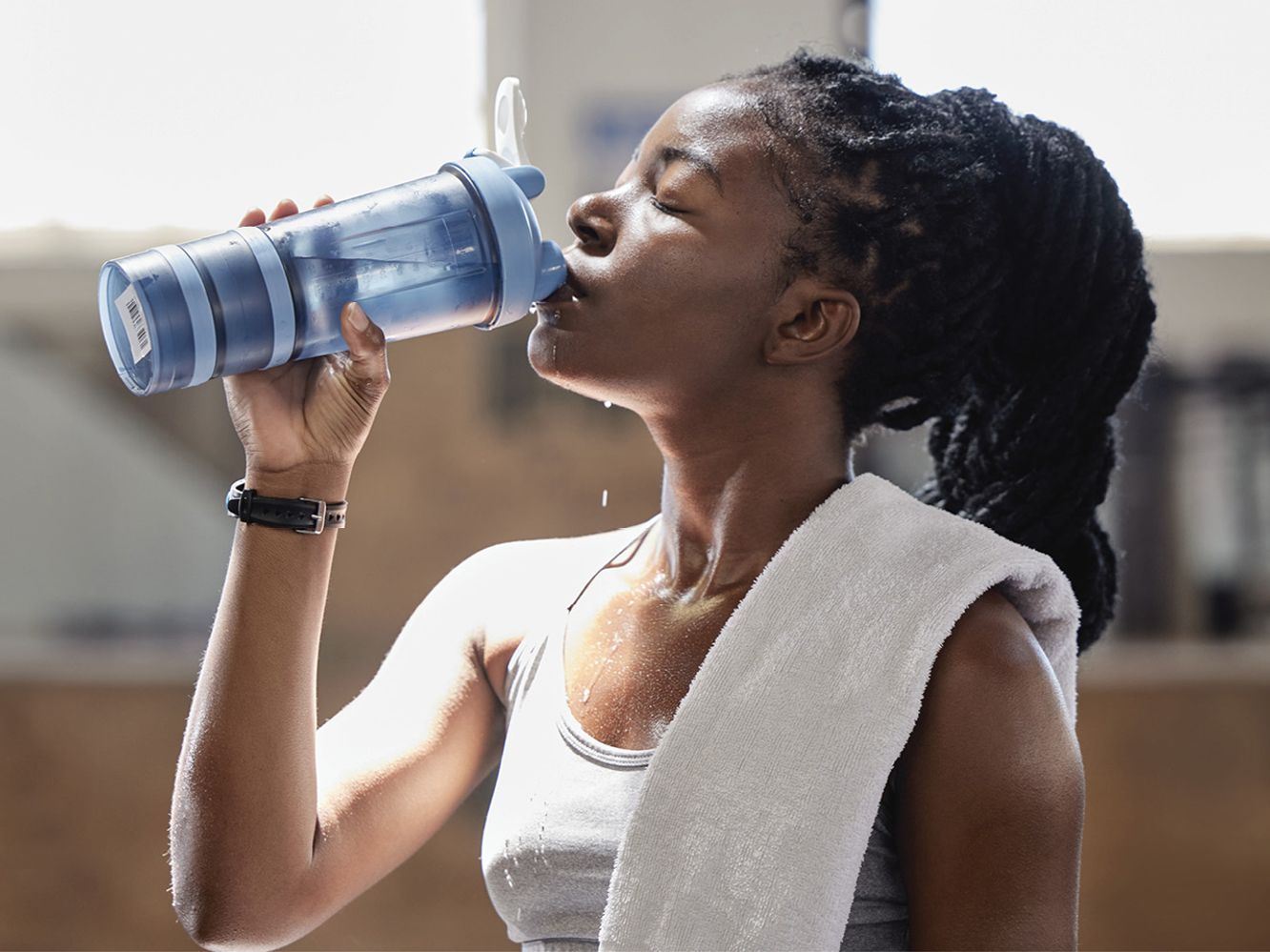 A woman in workout attire drinks from a light-blue water bottle, with a white towel on her left shoulder