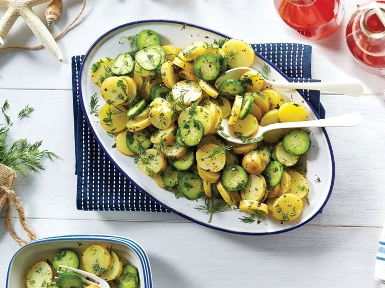 A cucumber and potato salad with fresh dill and parsley and fingerling potatoes on a white platter with a blue border with two white salad tongs resting in it.