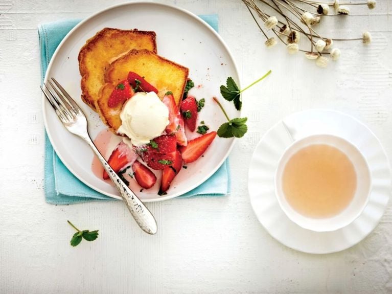 Cake toast with honeyed strawberries, made of Sara Lee store bought pound cake, topped with a scoop of vanilla ice cream on a white plate with a fork resting on it next to a cup of tea