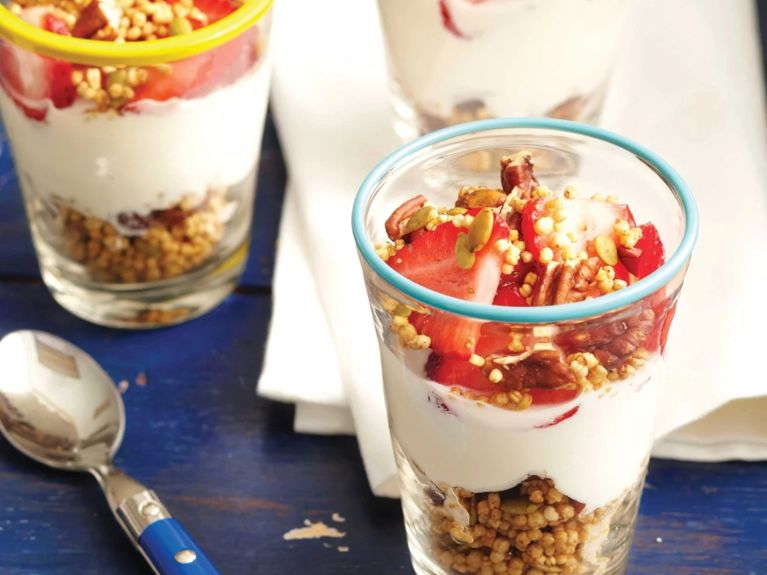 Gluten-free strawberry and granola parfaits topped with chopped pecans and puffed quinoa in tall glasses next to blue spoons