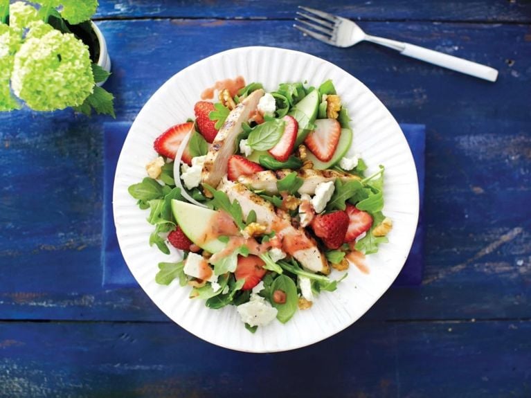Grilled chicken and strawberry salad with arugula and walnuts and apples and feta on a white plate next to a bouquet of green hydrangeas