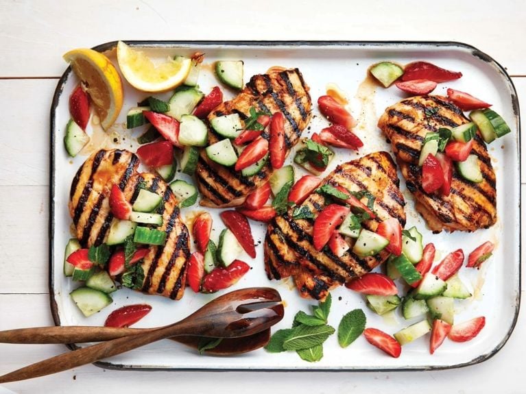 Spicy grilled chicken with strawberry-cucumber salad on a white platter with slices of lemon and wood tongs