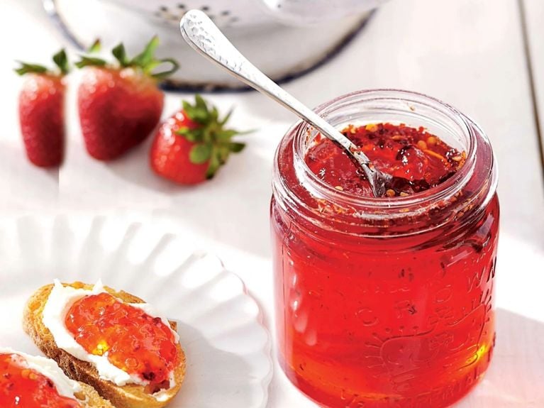 A jar of spicy strawberry jelly topped with red pepper flakes with a spoon resting in it next to toast topped with the jelly next to a white colander full of fresh strawberries