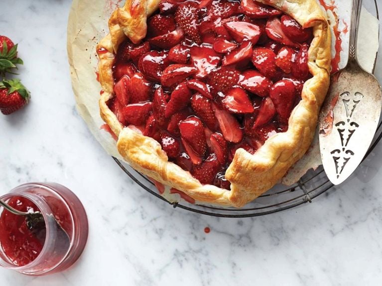 A strawberry galette—strawberries topping a rustic free-form pie crust—on a cooling rack next to a vintage cake server. The galette is on a marble counter, next to it are two fresh strawberries and a jar of strawberry jam with a spoon in it
