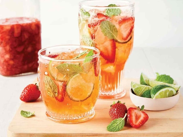 A mason jar filled with gin and strawberries to make strawberry gin next to a short and tall glass filled with strawberry fizz gin cocktail with ice, sliced strawberries, mint lives and slices of lime, next to sliced strawberries and lime on a wood cutting board on a wood table