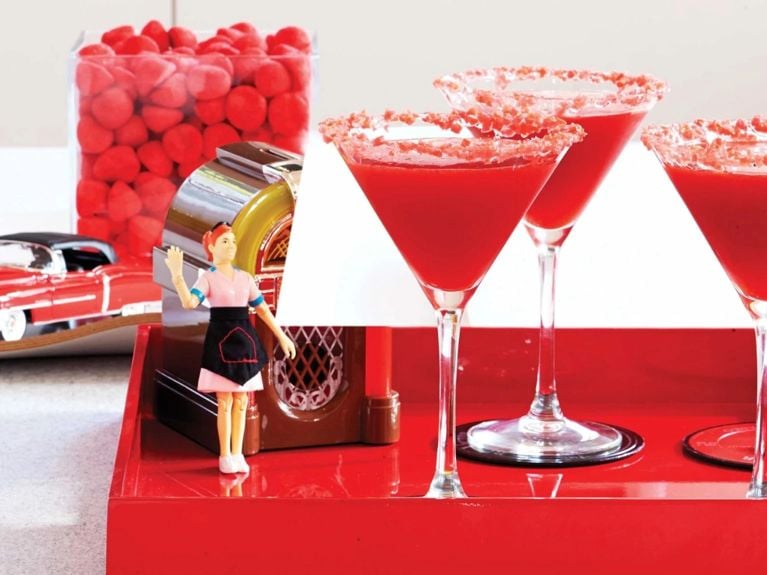 A strawberry pop rocks martini next to a toy jukebox and a toy waitress; the martini is in a martini glass and rimmed with red pop rocks; it's made with vanilla vodka; strawberry purée; vanilla vodka for a post on a strawberry pop rocks martini recipe