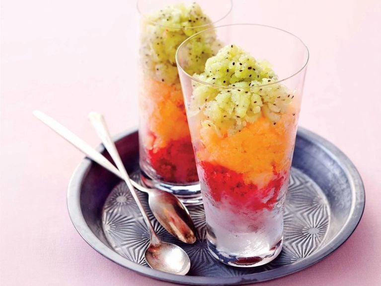 Two strawberry-rhubarb granitas on a tray, the drinks are layered with green kiwi granita on top then orange granita then the strawberryy rhubarb granita at the bottom with spoons next to them on a pink background