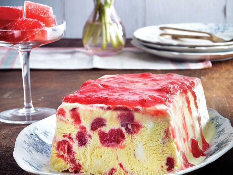 Strawberry rhubarb semifreddo on a blue and white plate next to a coupe glass filled with rhubarb gummies in front of a vase of pink tulips