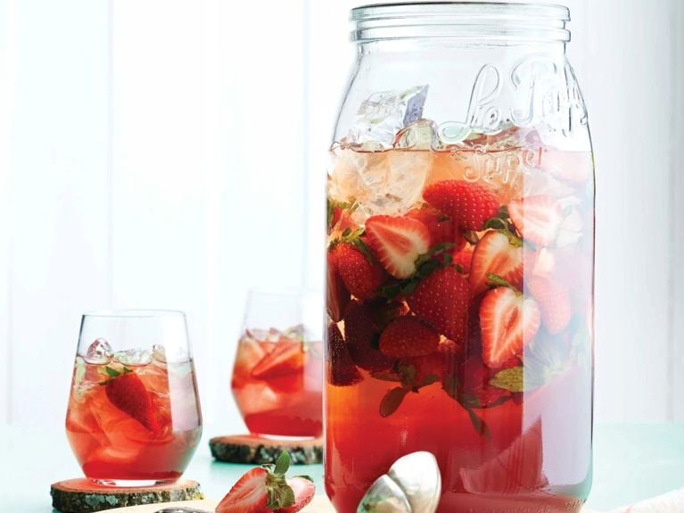A large mason jar filled with strawberry sangria with fresh strawberries floating in it, next to it is a vintage serving ladle and two stemless wine glasses filled with ice and sangria; all are on a pale blue table next to baby's breath and a checked linen tea towel