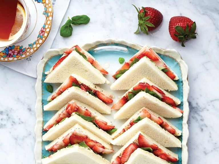 Strawberry tea sandwiches—crustless white bread sandwiches with strawberries and fresh basil—on a blue ceramic tray edged with gold and white scalloped edges on a marble surface next to a cup of tea in a floral cup and saucer with a slice of lemon on a white lace napkin next to fresh strawberries