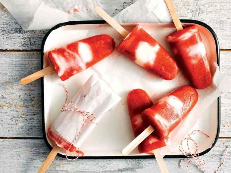 Strawberry-vanilla yogurt ice pops—which are red with white stripes—on a white tray. One is wrapped with parchment paper and red and white string
